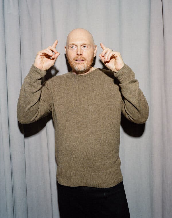 Bill Burr, in a taupe sweater and black pants stands with his hands raised and his index fingers pointing at his own head.